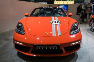 Front view of an orange Porsche 718 convertible featuring racing stripes, a 718 hood emblem, and a sporty design at an indoor car exhibition.