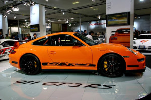 Orange Porsche 911 GT3 RS sports car shown from the side on an indoor exhibition floor.